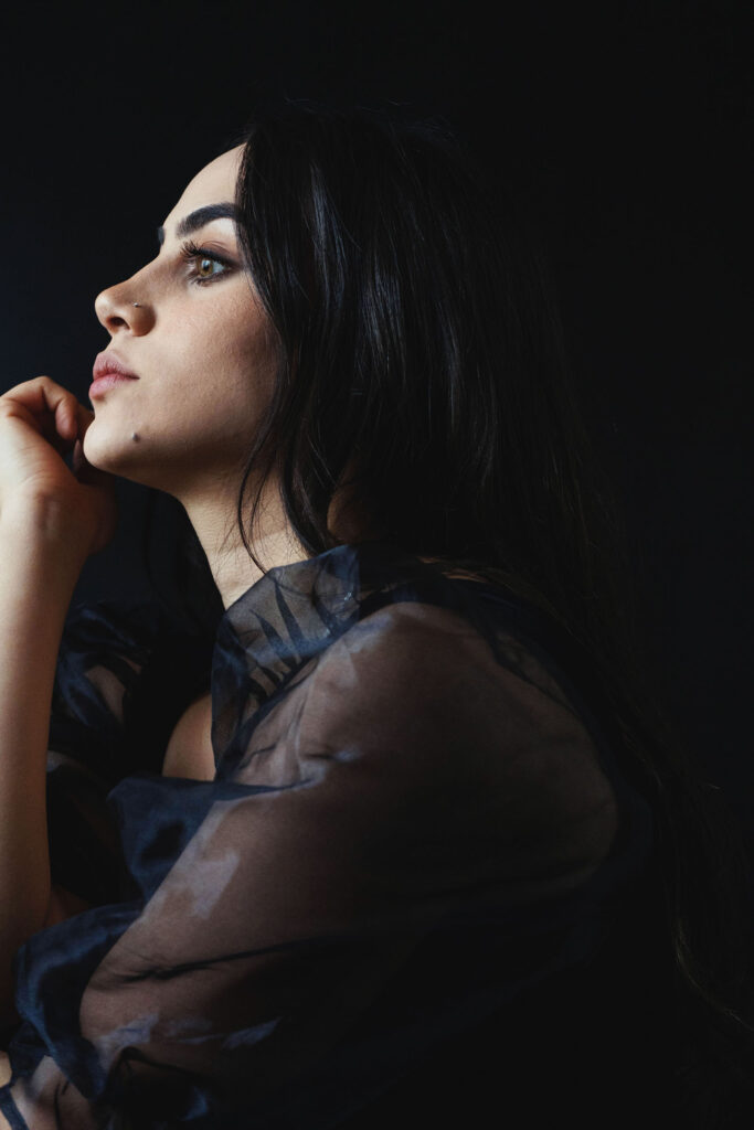 Valeria Cisneros a model with striking features is captured in a side profile against a dark backdrop. She rests her chin on her hand her gaze directed upwards exuding a sense of contemplation and elegance.